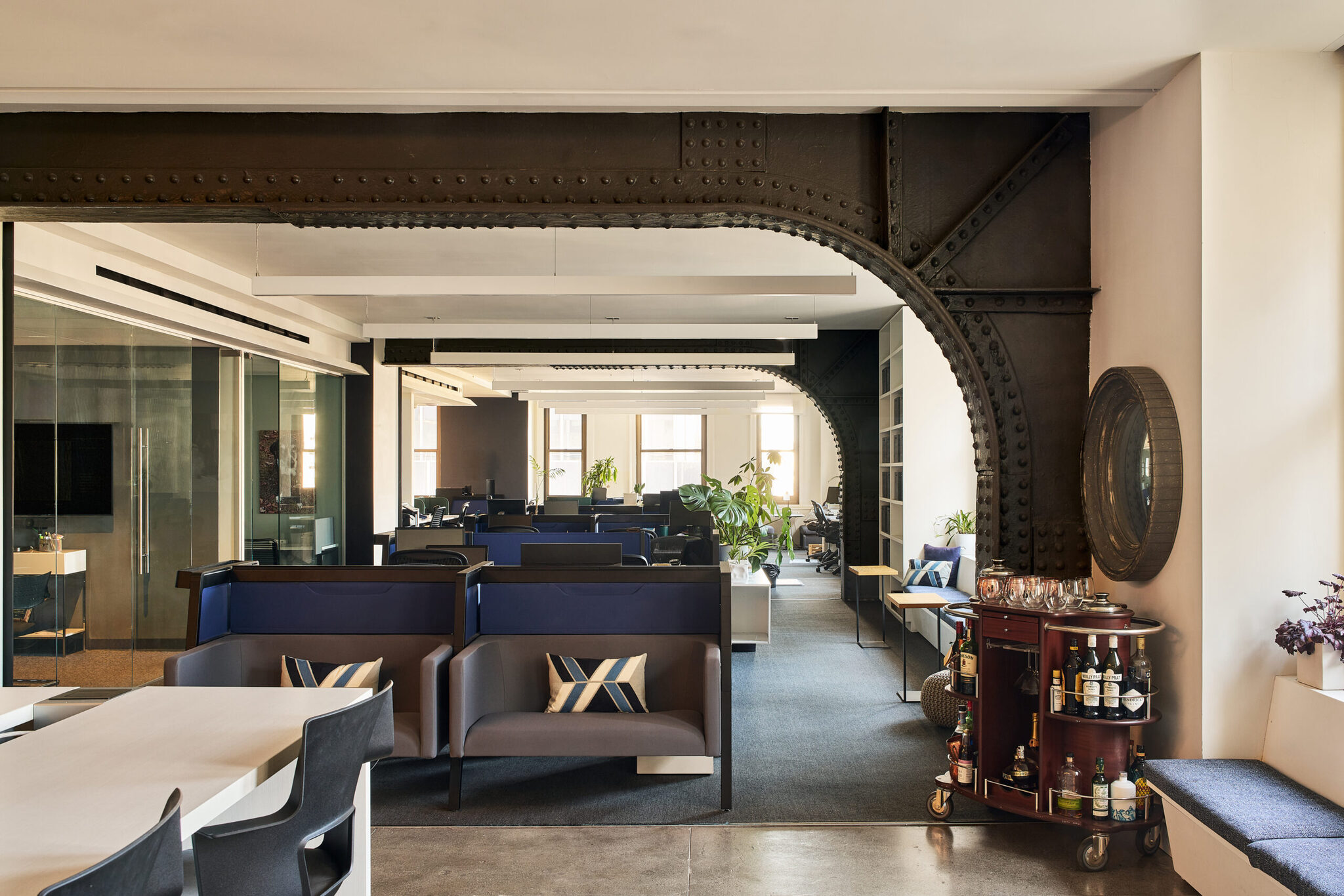 Workplace lounge framed by an exposed riveted steel arch, with blue banquettes and a bar cart